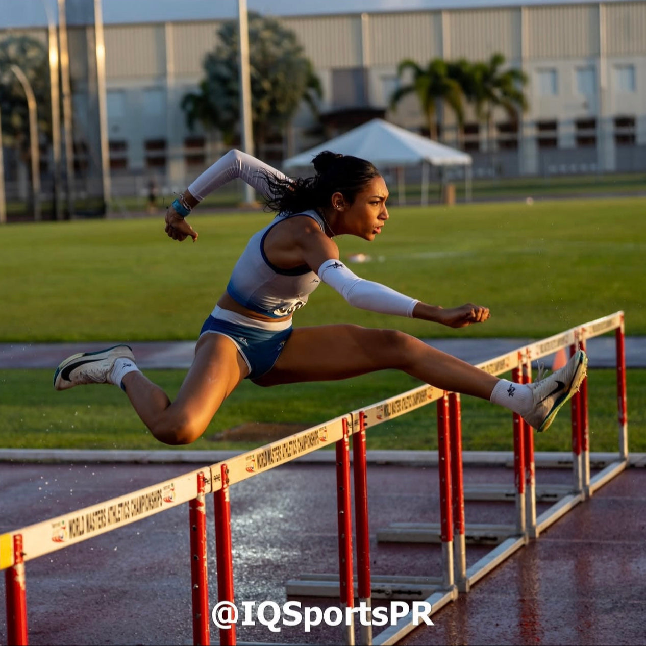 Athlete jumping over hurdles on a track with a building and trees in the background